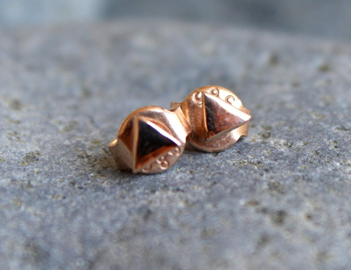 The backs of two rose gold triangle stud earrings with butterfly clasps, resting on a gray textured surface.