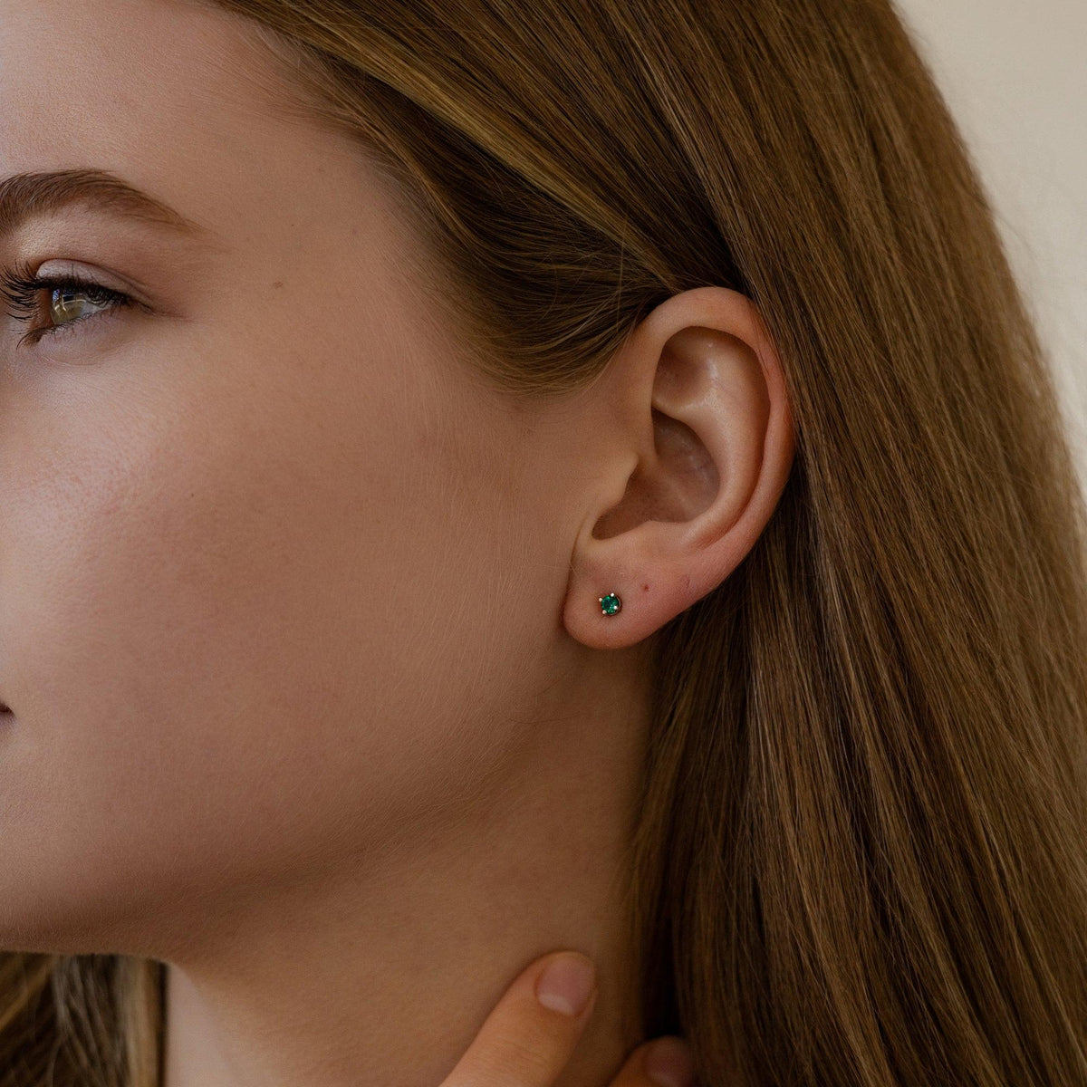A close-up of a woman's ear, showing a small, square-cut emerald stud earring in a silver-toned setting on her lobe.
