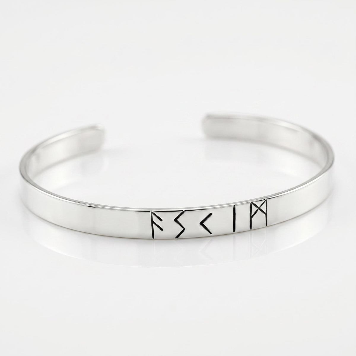 A close-up studio shot of a smooth silver-colored metal cuff bracelet featuring several engraved rune characters on a reflective white surface.