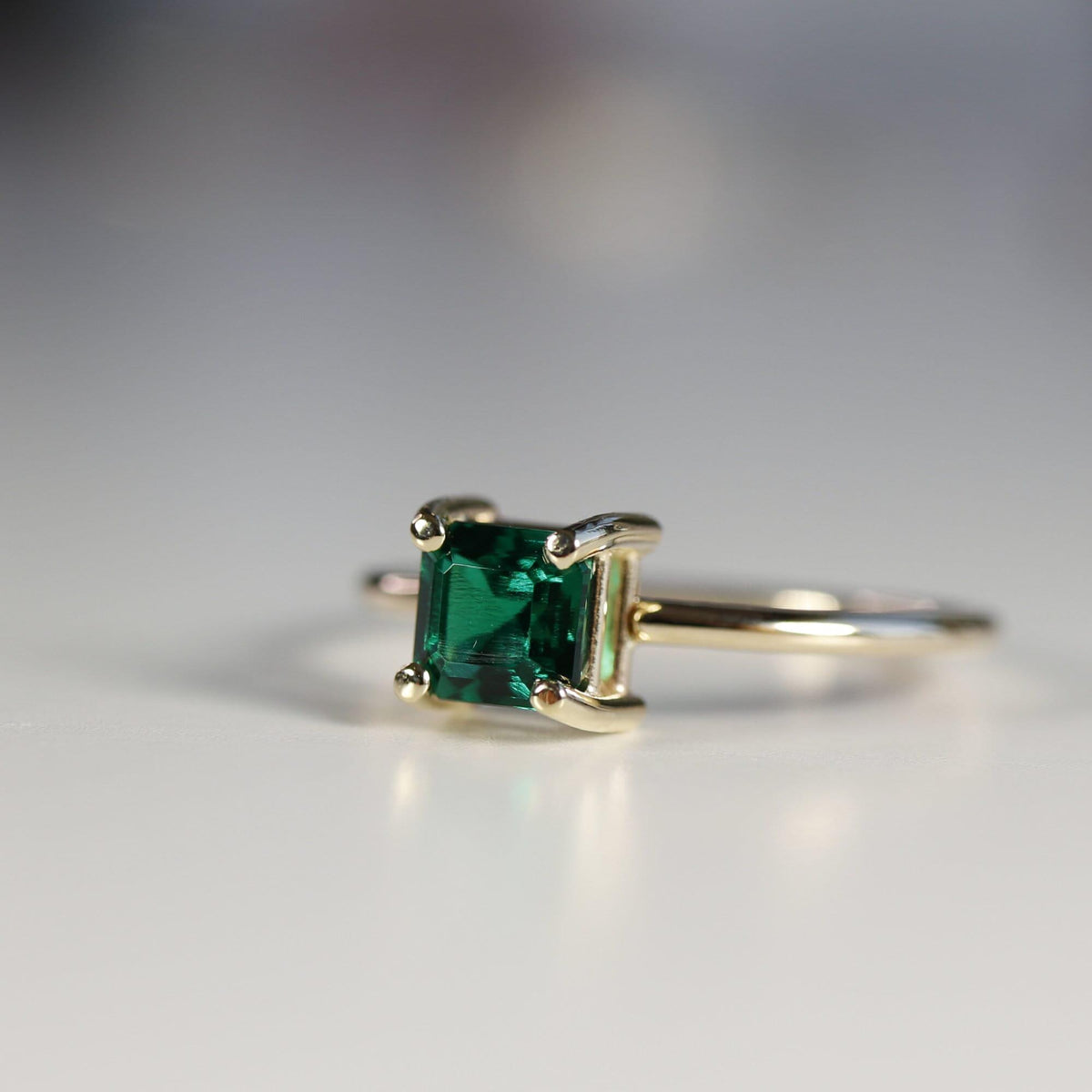 Side profile of a gold ring featuring a prong-set square green gemstone resting on a white surface.
