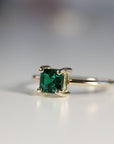 Side profile of a gold ring featuring a prong-set square green gemstone resting on a white surface.