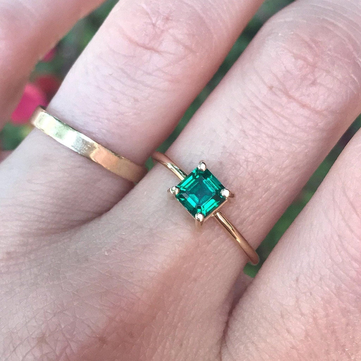 Close-up of a hand wearing a gold ring with a square green gemstone, accompanied by a plain gold band.