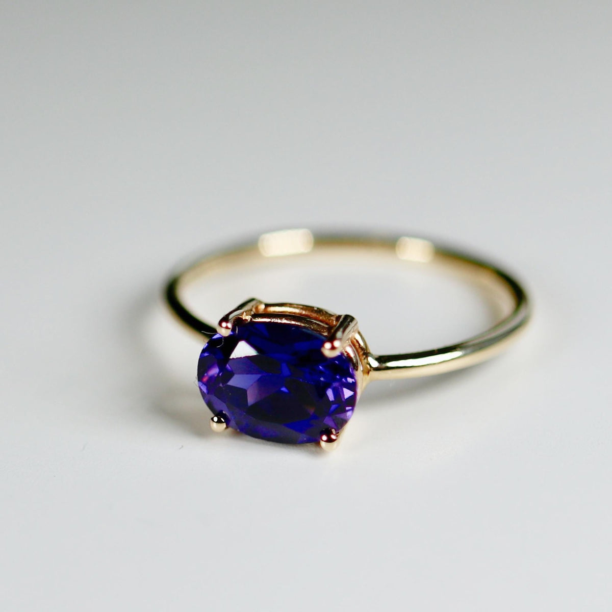 Angle shot of a gold ring with a prong-set oval purple gemstone on a clean white surface.