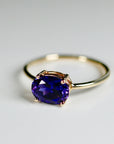 Angle shot of a gold ring with a prong-set oval purple gemstone on a clean white surface.