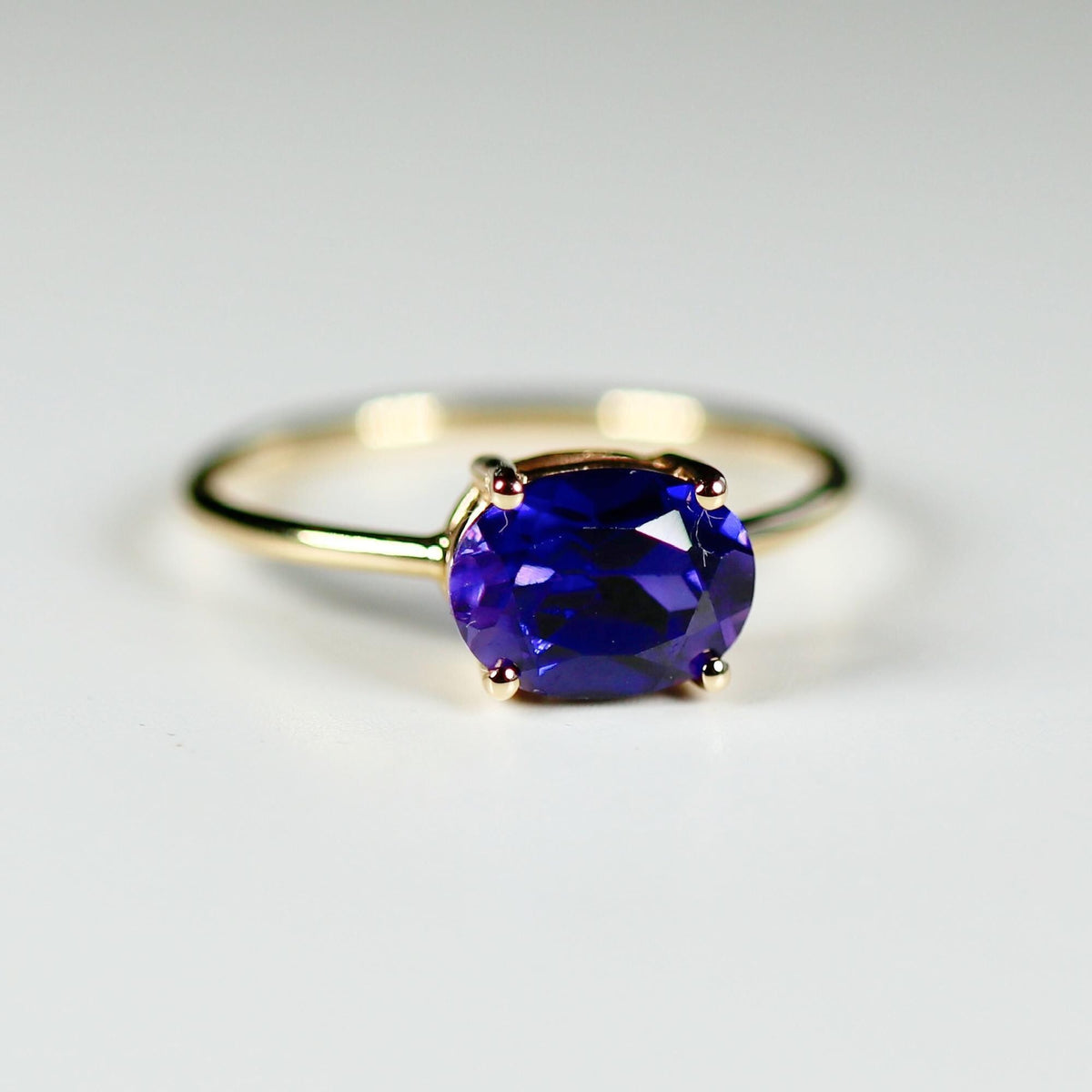 Slightly angled macro view of a gold ring holding an east-west set oval purple faceted gemstone on a white background.