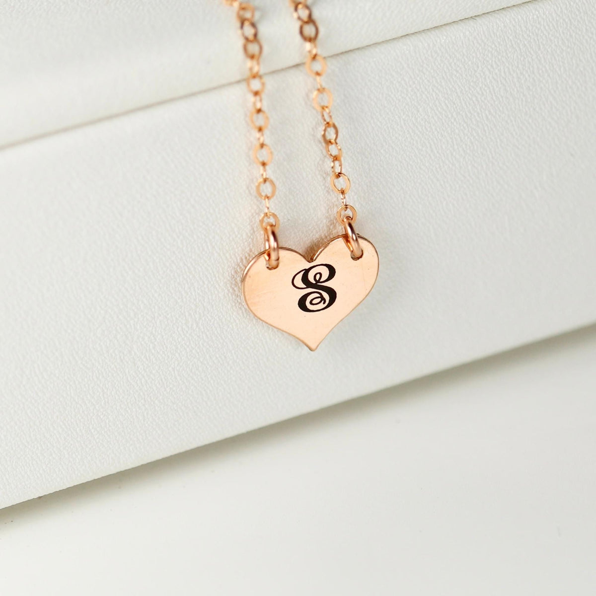 A rose gold necklace with a heart pendant engraved with the letter S rests against a white background.
