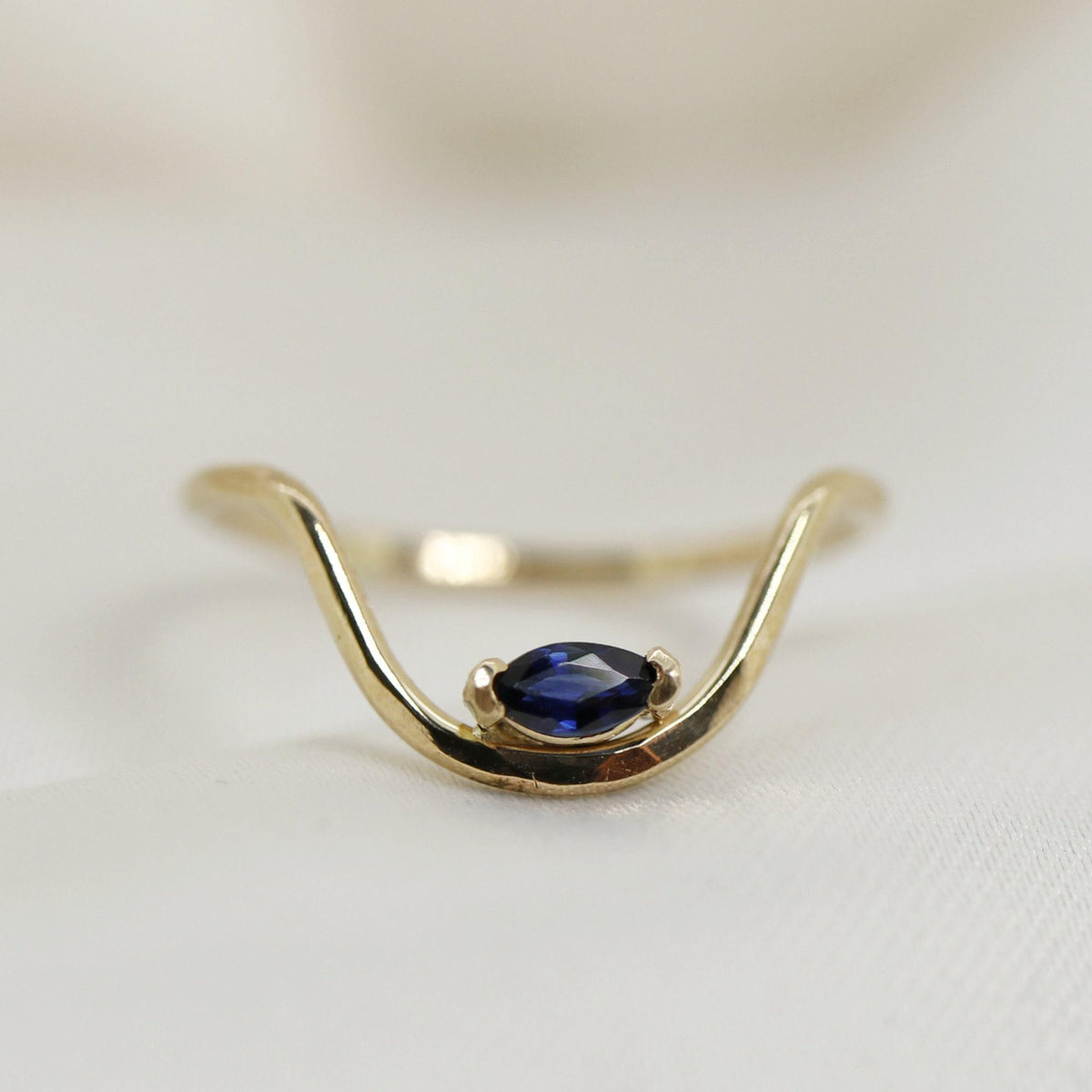 A curved gold ring featuring a single dark blue marquise-cut sapphire set horizontally in the center, resting against a soft white textured surface.