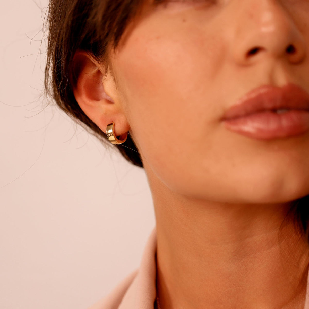 A woman's ear wears a thick gold hoop earring with a small diamond set in a starburst pattern on the front.