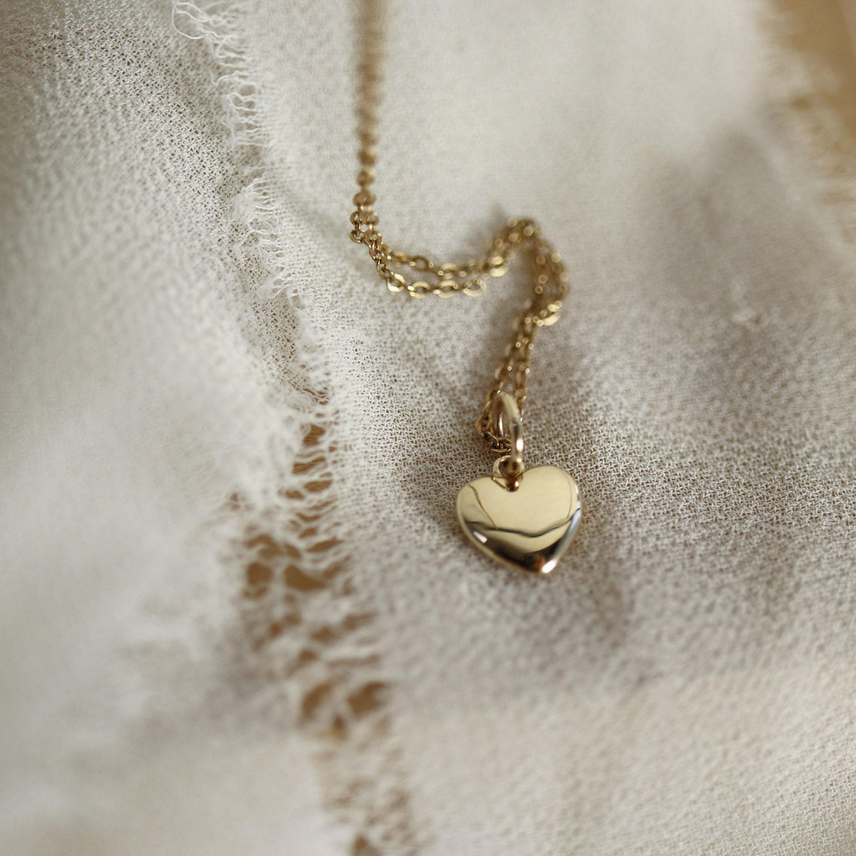 A small, shiny gold heart pendant on a delicate gold chain is partially visible on a textured white fabric.