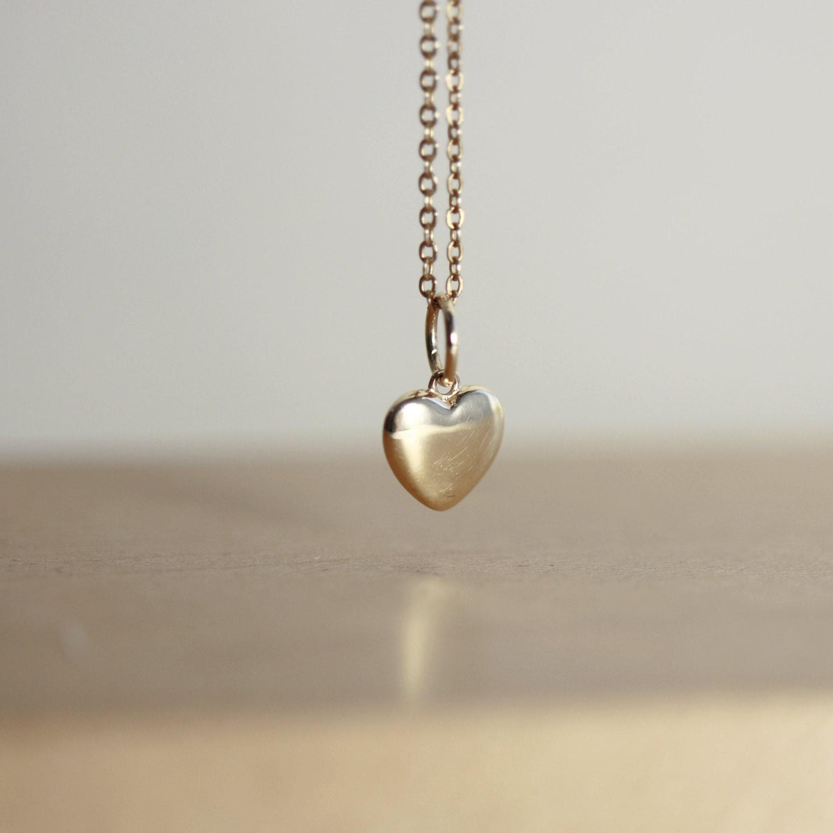 A small, shiny gold heart pendant with a bail hangs from a delicate gold chain, centered against a blurred light brown background.