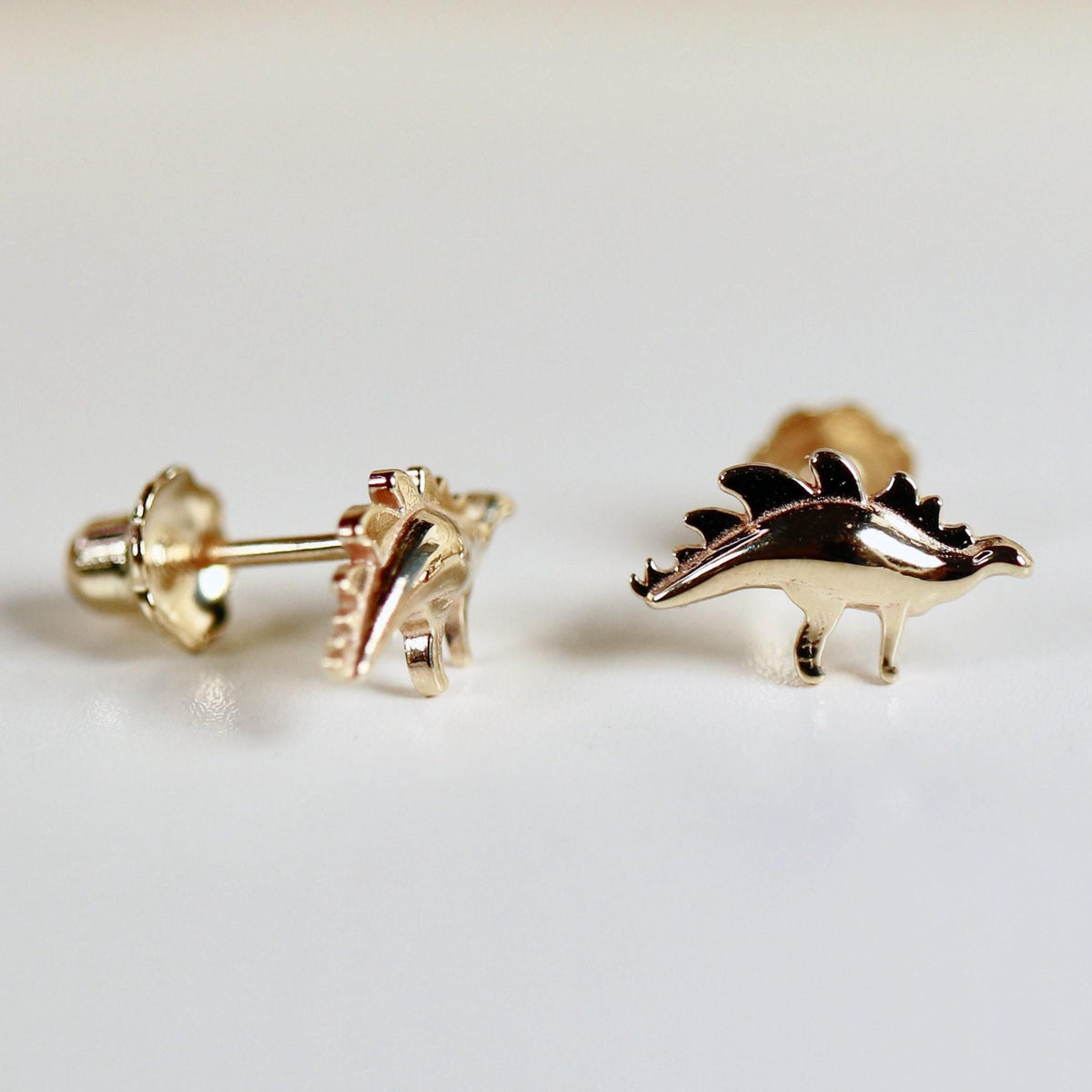 Two shiny gold dinosaur stud earrings are shown, one with its post and screw-back visible, the other facing forward.