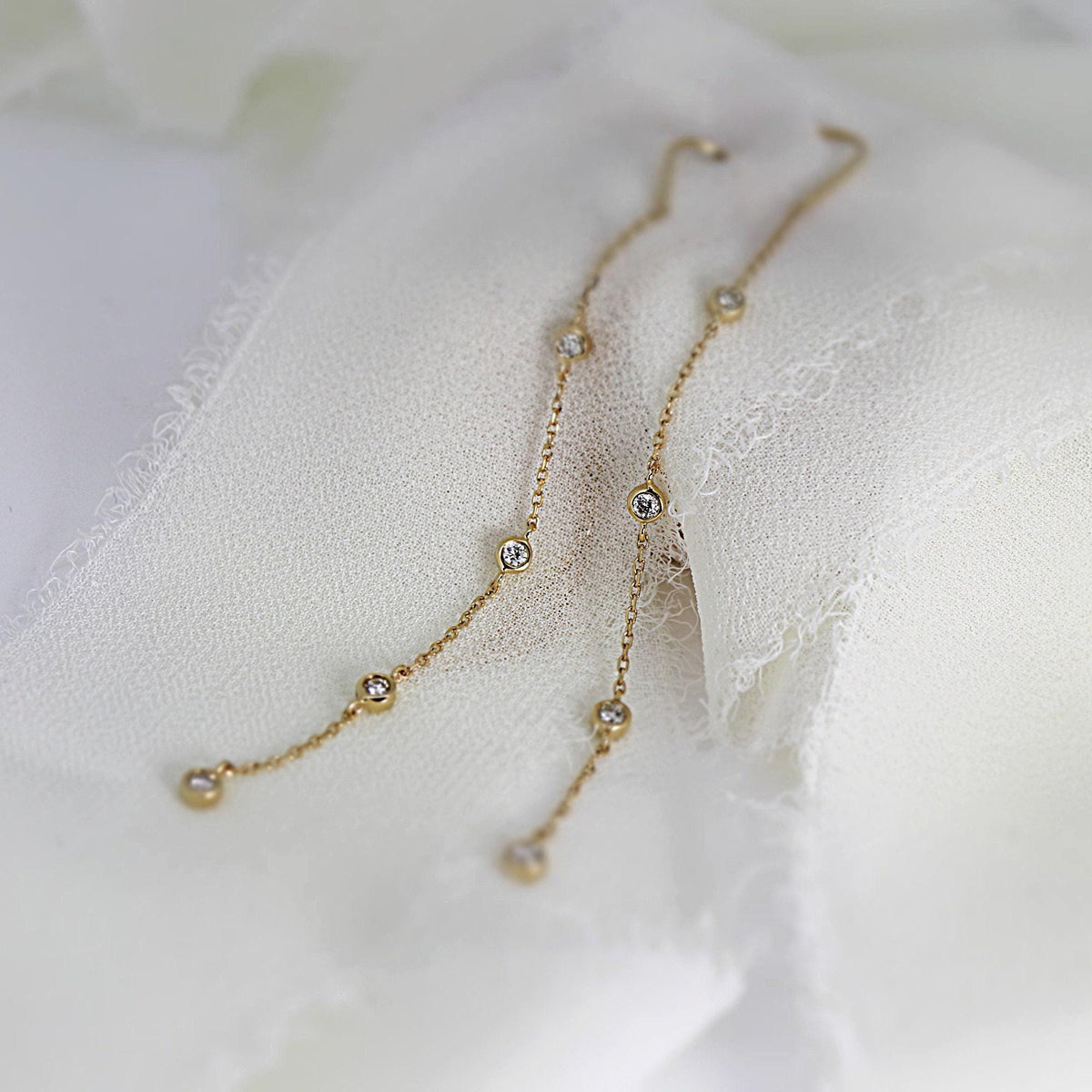 Two gold chain threader earrings with diamond stations resting on white crumpled fabric.