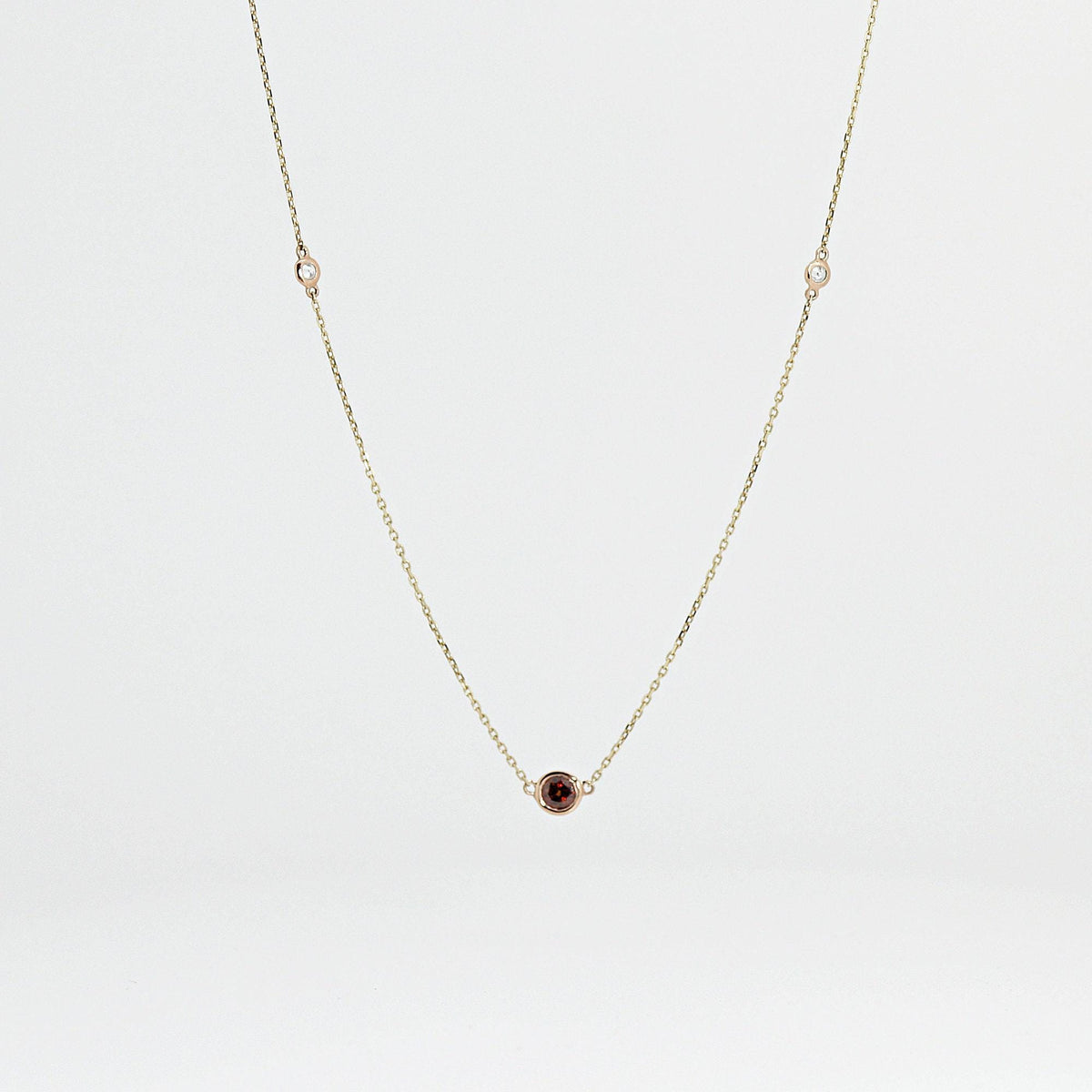 A gold chain necklace with a single red oval gem in the center and two clear round gems spaced evenly on either side, all set in bezels, is displayed against a light gray background.