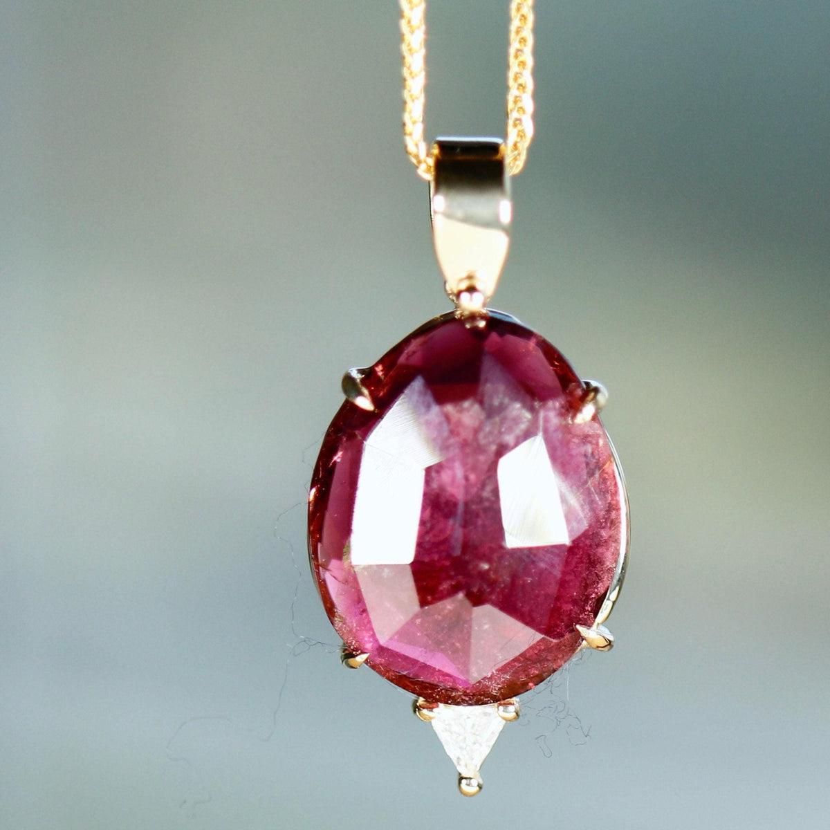 A close-up of a gold necklace with an oval, faceted, dark pink tourmaline pendant, with a small triangular diamond set below it, on a light gray background.