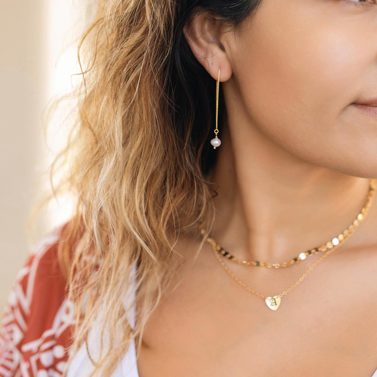 A person wears a layered gold necklace, with a choker of flat gold discs and a longer chain with a small heart pendant engraved with a white letter &amp;quot;A,&amp;quot; and a gold earring with a pearl.