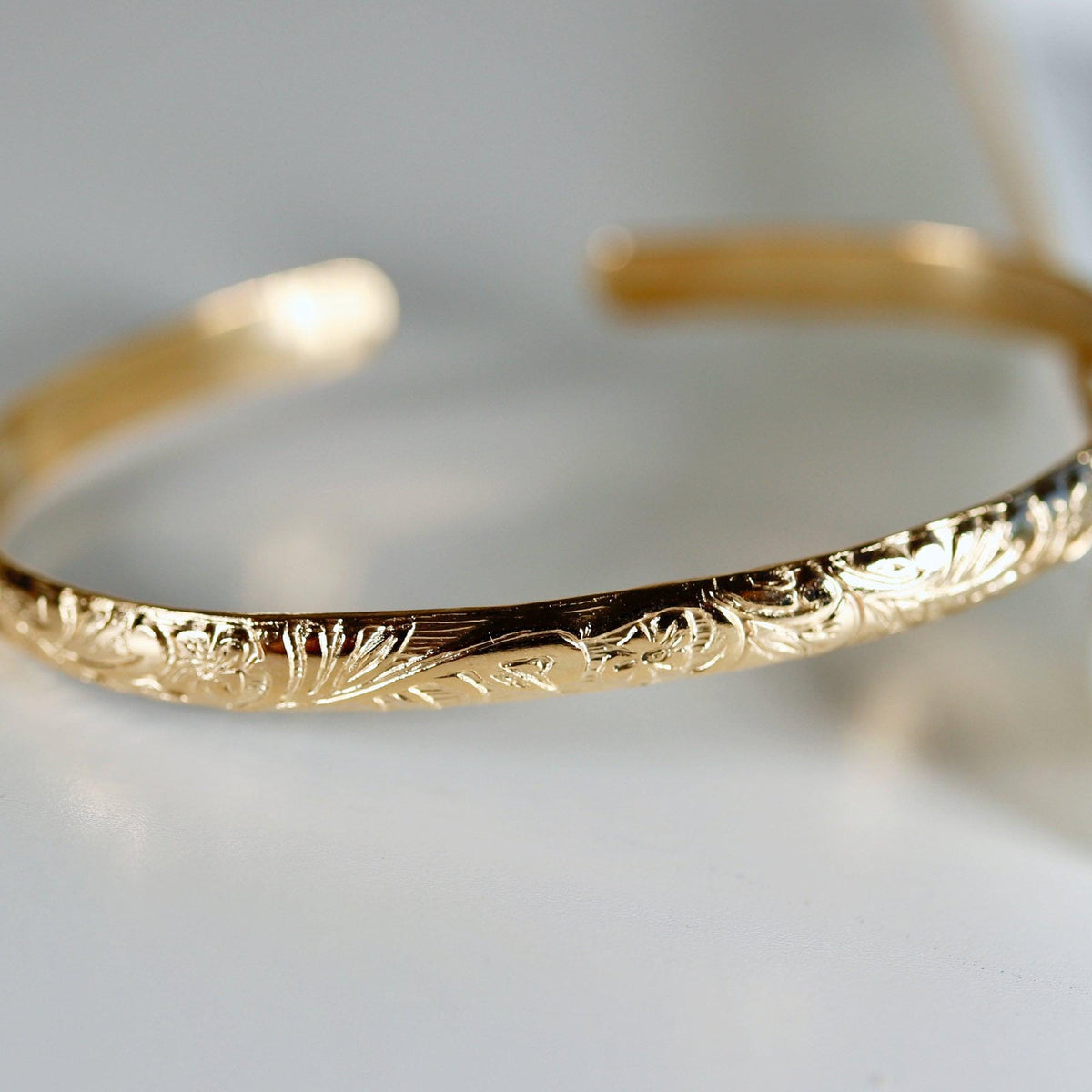 A gold cuff bracelet with a floral pattern engraved on its outer surface, shown on a white background.