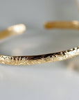 A gold cuff bracelet with a floral pattern engraved on its outer surface, shown on a white background.