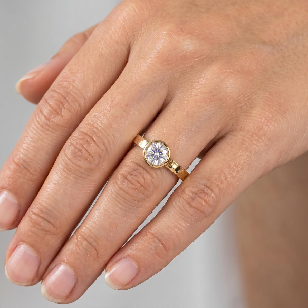 A person's hand wears a gold ring with a hammered band and a round, bezel-set diamond.