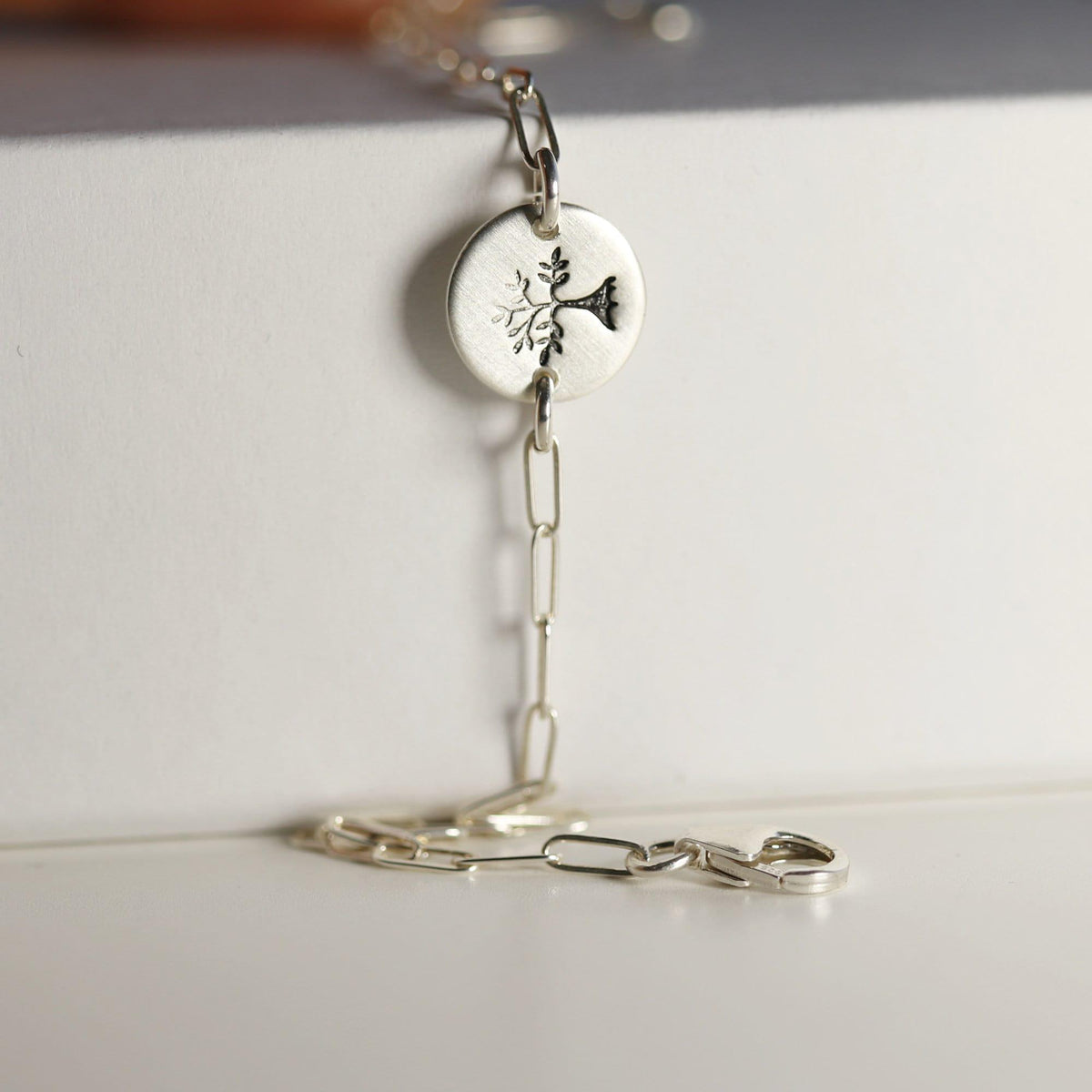 A silver paperclip chain bracelet with a round, flat silver charm featuring a stamped tree design, resting on a white surface.