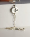 A silver paperclip chain bracelet with a round, flat silver charm featuring a stamped tree design, resting on a white surface.