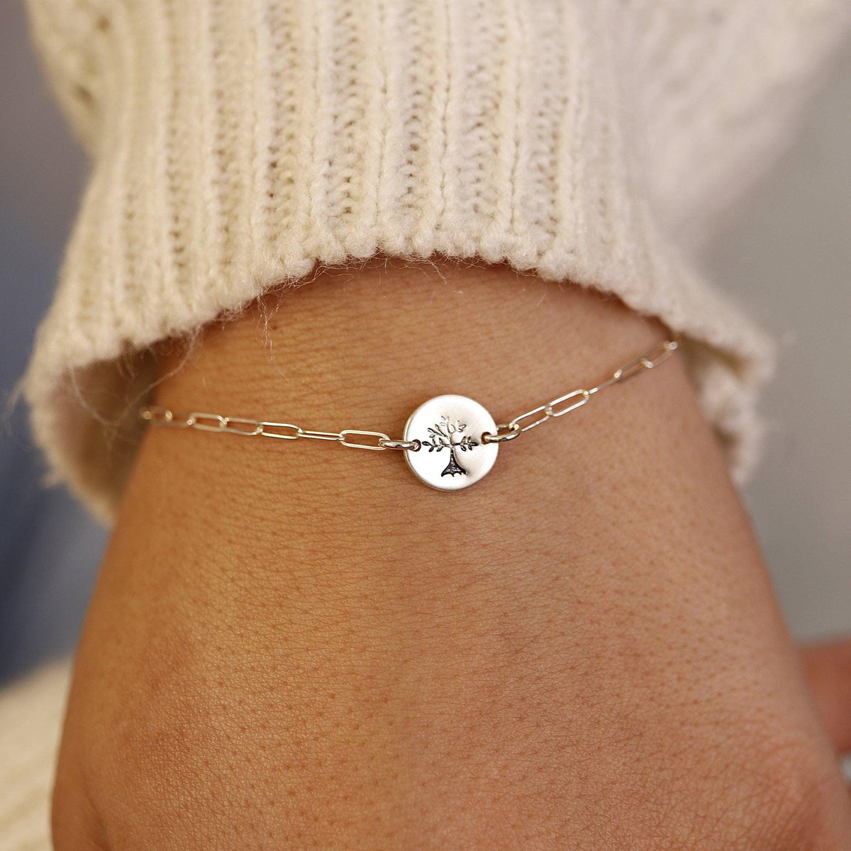 A silver paperclip chain bracelet with a round, flat silver charm featuring a stamped tree design, worn on a wrist with a cream-colored sweater sleeve visible.