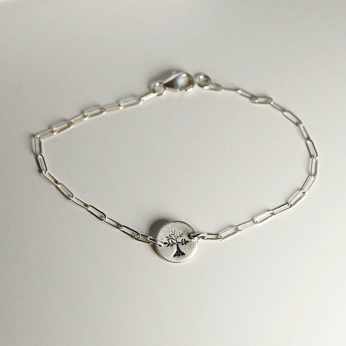 A silver paperclip chain bracelet with a round, flat silver charm featuring a stamped tree design, laid flat on a white surface with the clasp visible.
