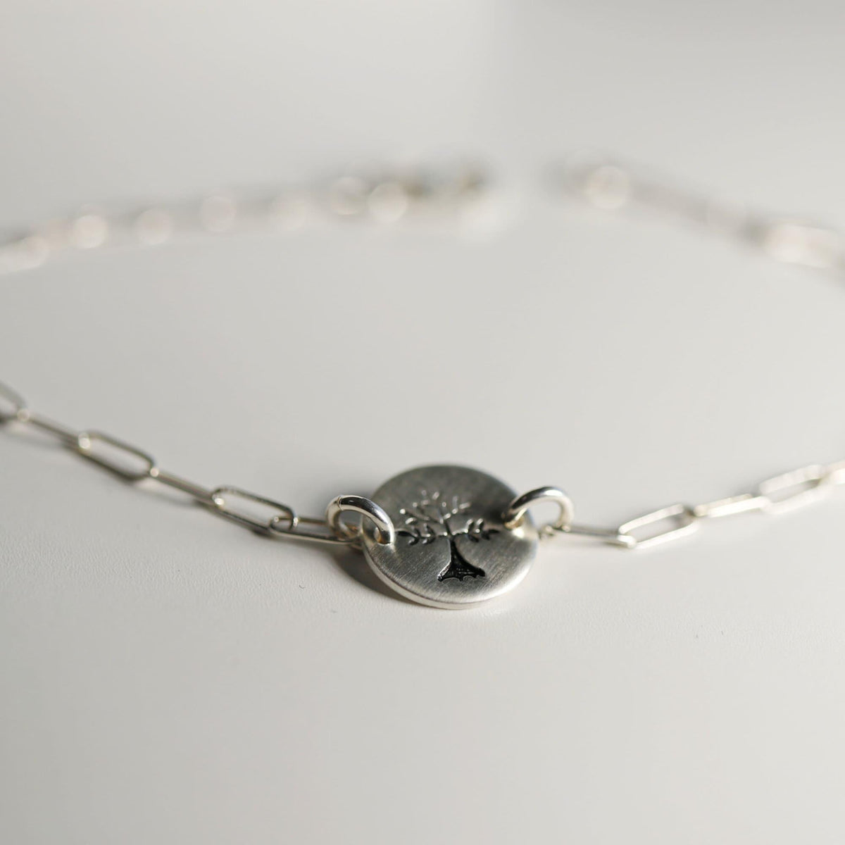 A close-up, slightly blurry shot of the round, flat silver charm with a stamped tree design, attached to a silver paperclip chain bracelet on a white surface.
