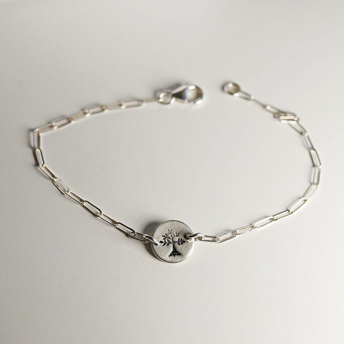 A silver paperclip chain bracelet with a round, flat silver charm featuring a stamped tree design, laid flat on a white surface, showing the full length of the bracelet.