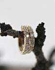 A single gold huggie hoop earring, encrusted with two rows of diamonds, is resting on a dark, textured tree branch.