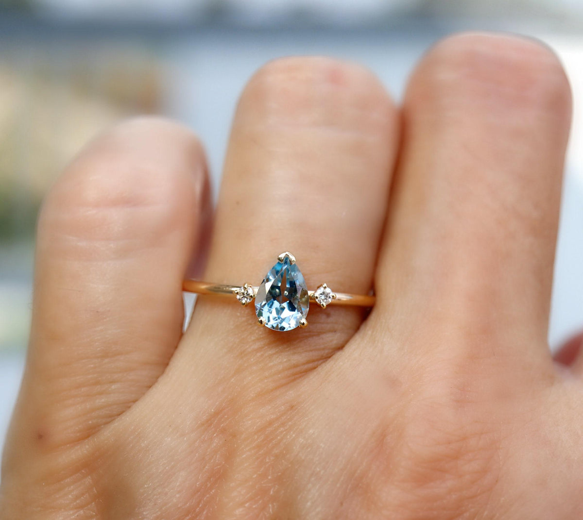 A hand models a gold ring with a pear-shaped aquamarine stone flanked by two small round diamonds.
