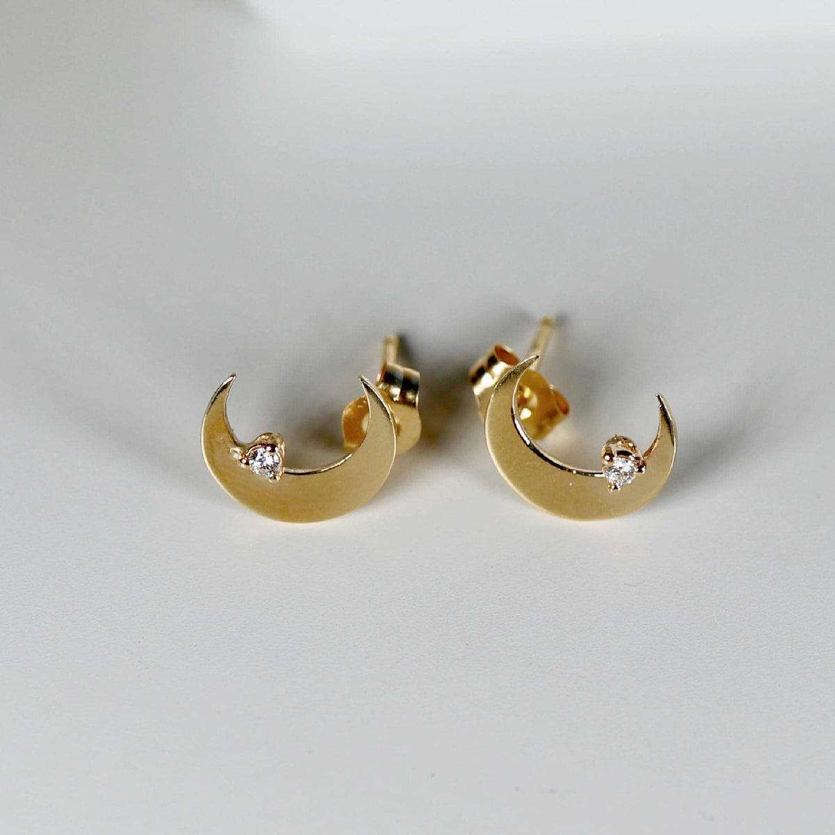 Two gold crescent moon stud earrings, each with a small clear stone set near the bottom curve, are displayed on a white surface.