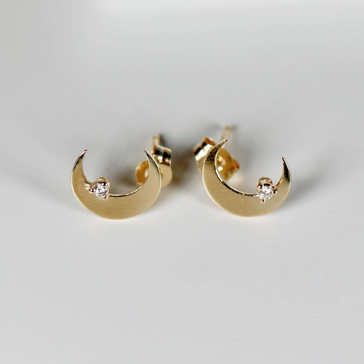 Two gold crescent moon stud earrings, each with a small clear stone, are shown from a slightly different angle on a white background.