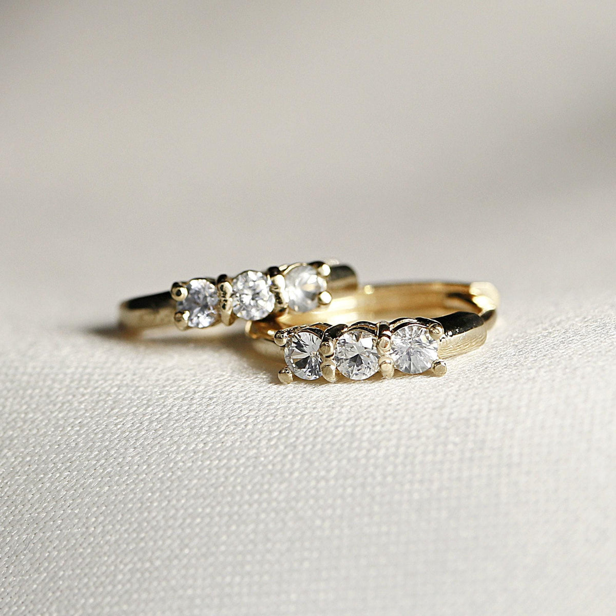 Two small gold huggie hoop earrings, each with three clear round stones, resting on a white textured fabric.