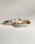 Two small gold huggie hoop earrings, each with three clear round stones, resting on a white textured fabric.