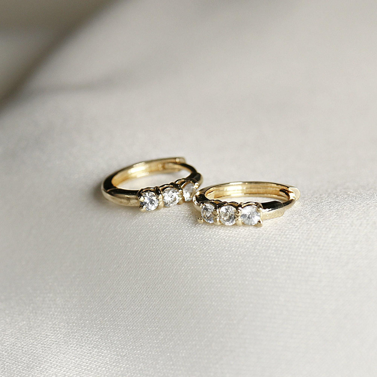 Two small gold huggie hoop earrings, each with three clear round stones, on a white satin-like fabric.