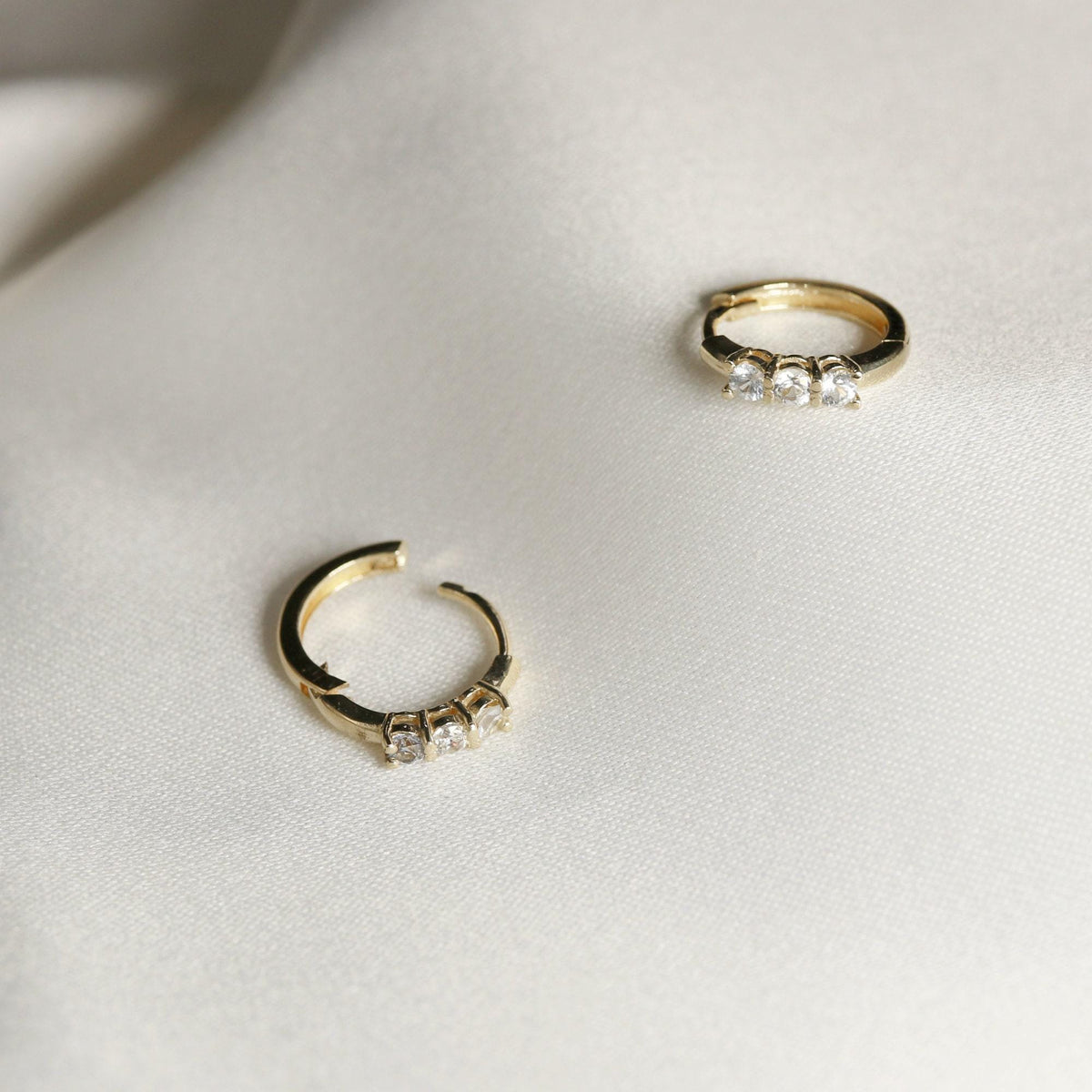 One small gold huggie hoop earring with three clear round stones, and one open gold huggie hoop earring with three clear round stones, on a white textured fabric.