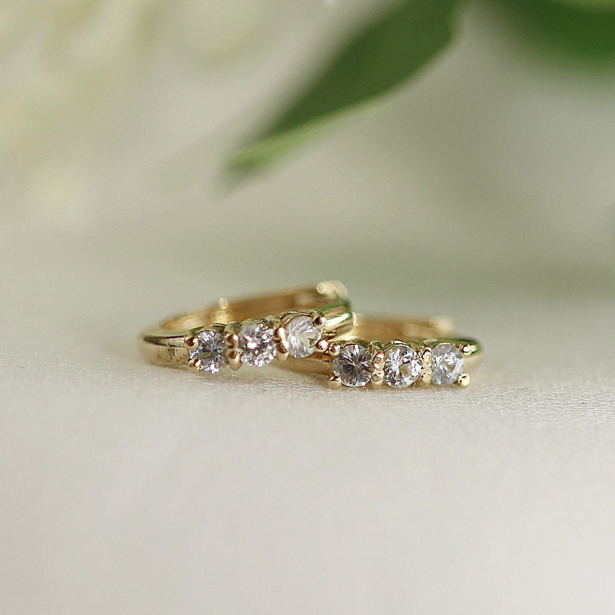 Two small gold huggie hoop earrings, each with three clear round stones, on a white surface with a blurred green leaf in the background.