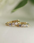 Two small gold huggie hoop earrings, each with three clear round stones, on a white surface with a blurred green leaf in the background.
