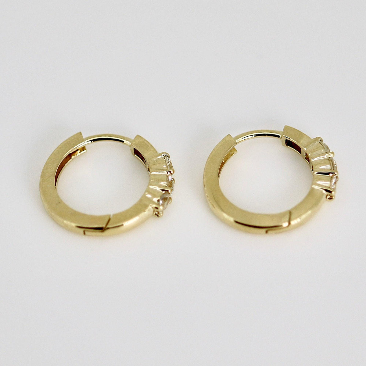 Two small gold huggie hoop earrings, each with three clear round stones on the front, viewed from the side on a light gray background.