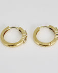 Two small gold huggie hoop earrings, each with three clear round stones on the front, viewed from the side on a light gray background.
