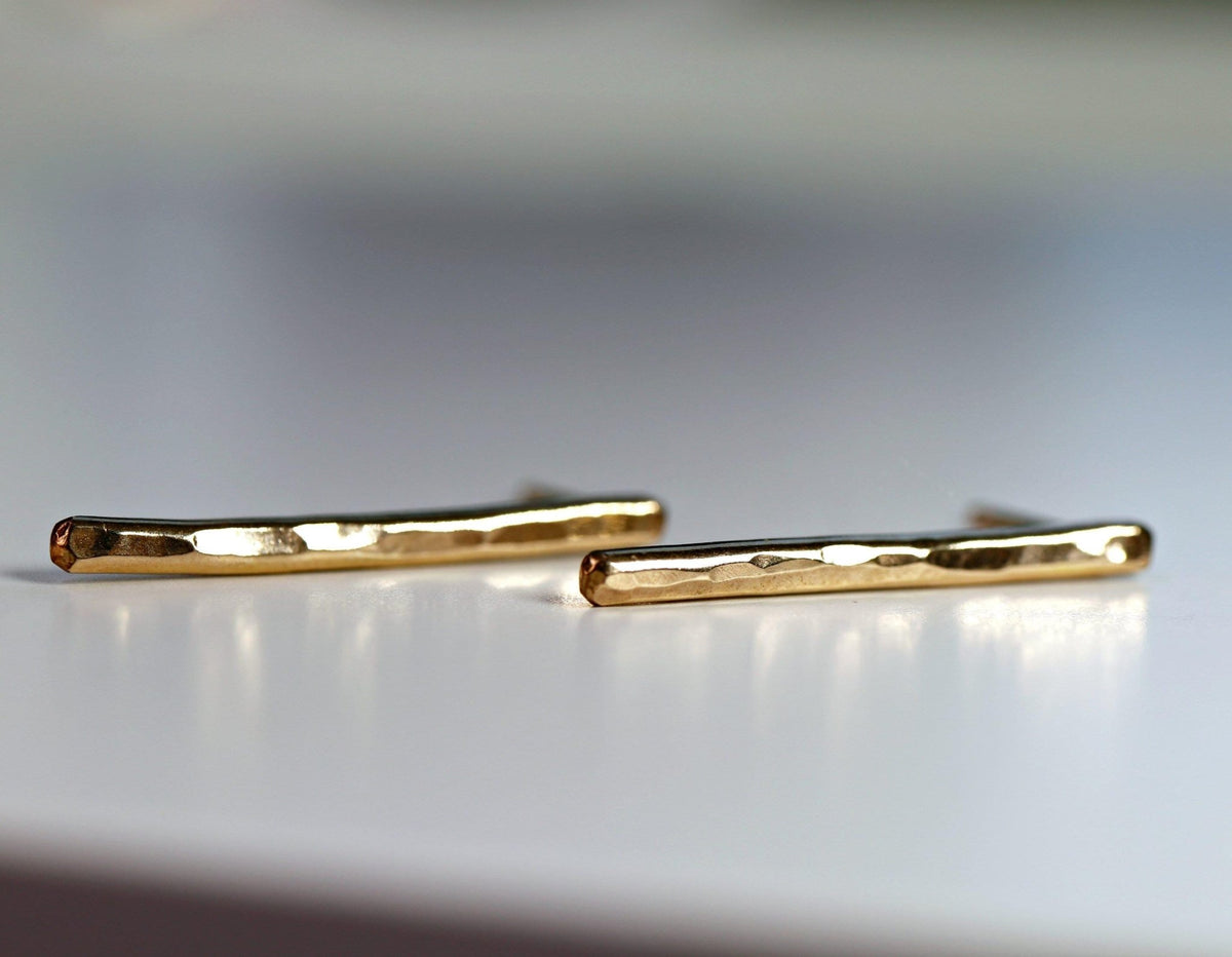 Two hammered gold bar earrings, one slightly in front of the other, lie on a white surface.