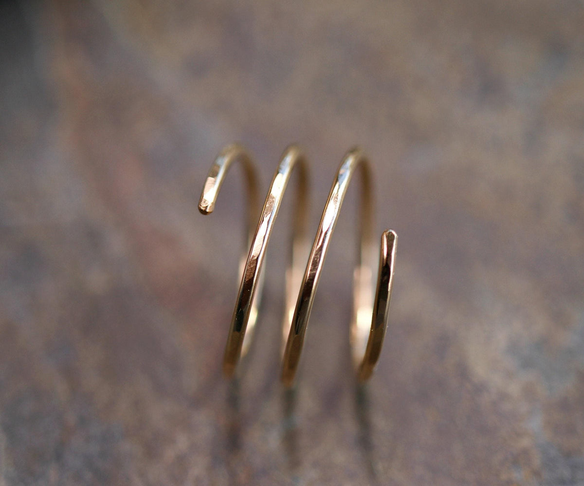 A gold-colored, three-coil spiral ring with hammered texture is shown on a blurred dark brown background.