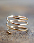 A gold-colored, three-coil spiral ring with hammered texture is shown on a blurred textured gray surface.