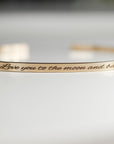 A close-up of a gold-colored open cuff bracelet shows the engraved text "Love you to the moon and back" across its surface.