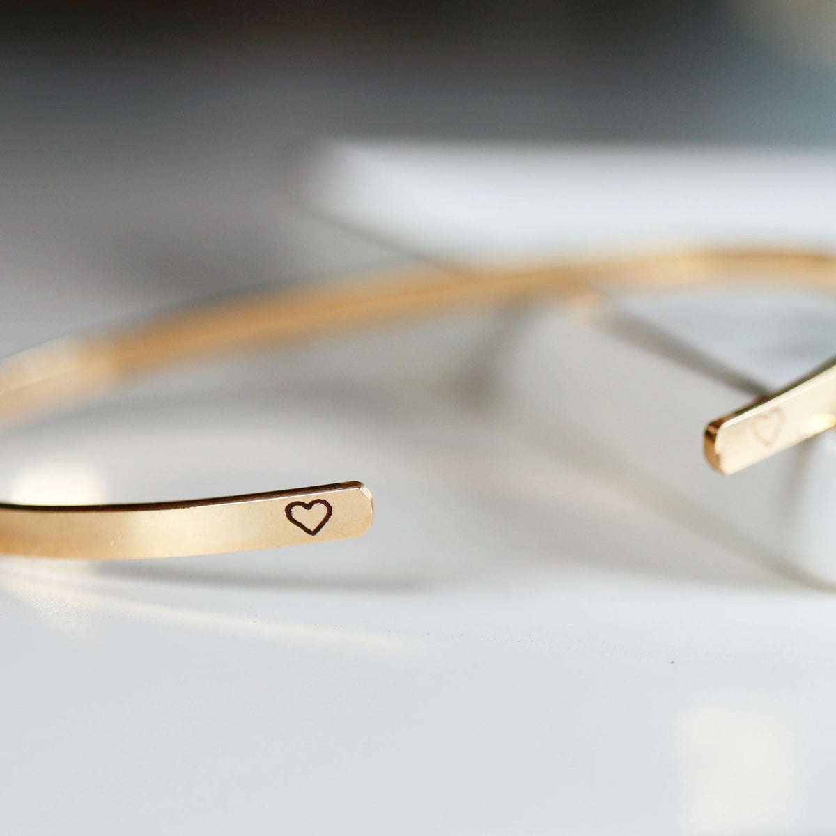 Two gold-colored open cuff bracelets are shown, each with a small engraved heart symbol on one end.