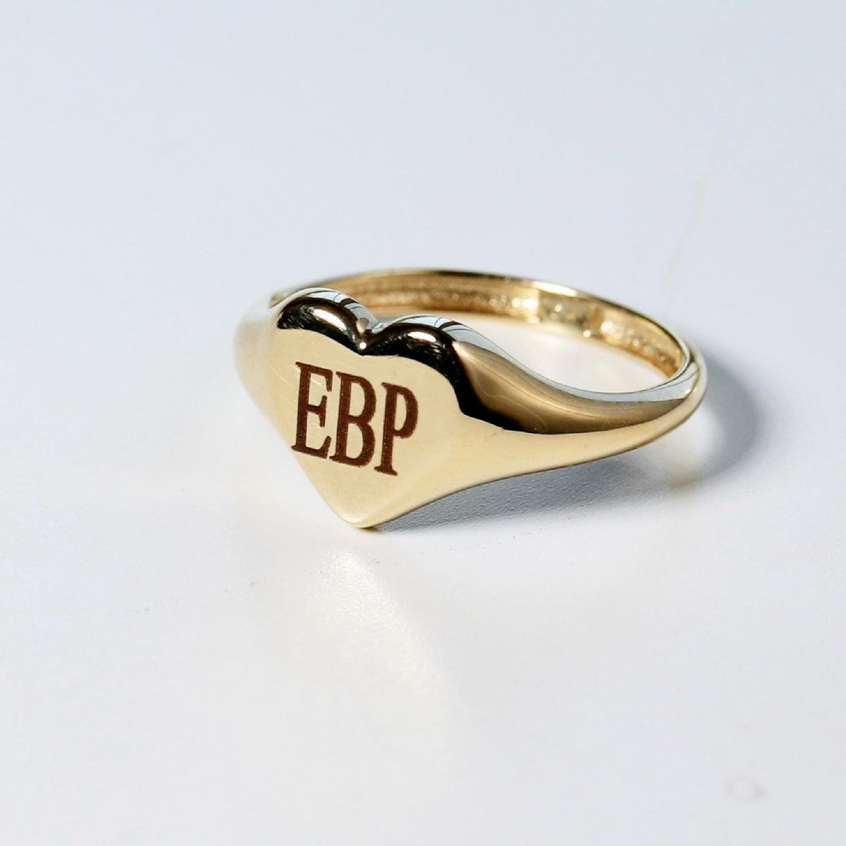 A gold heart-shaped signet ring with the initials "EBP" engraved on its face, sitting on a white surface.
