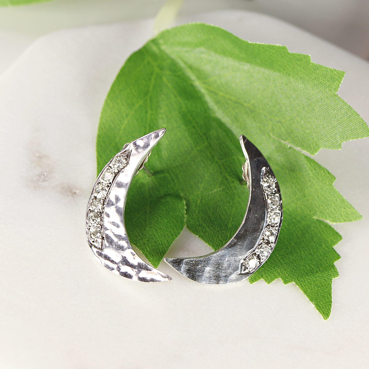 Two silver crescent moon earrings with stones placed on top of a green leaf against a white background.