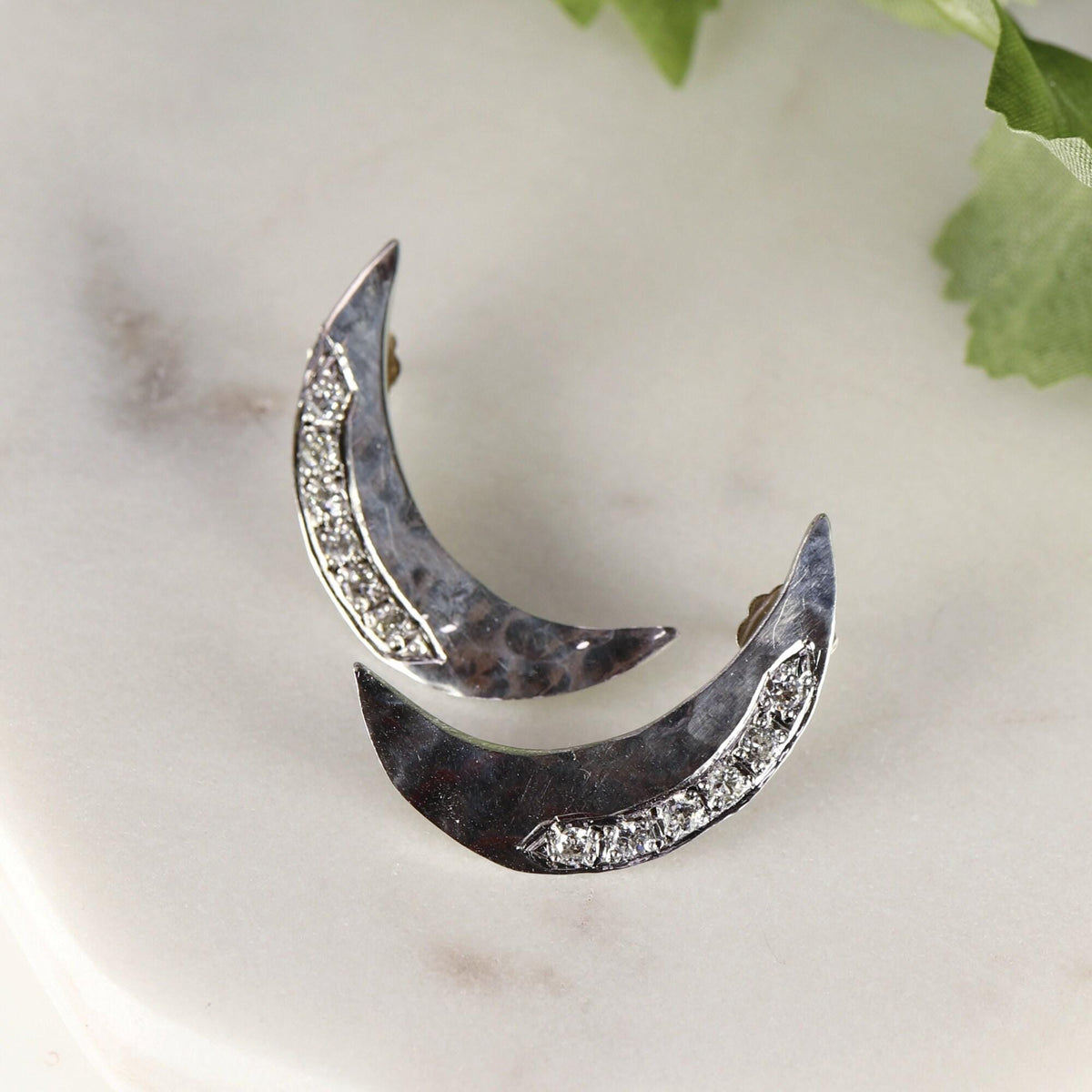 Two hammered silver crescent moon earrings with pave-set stones positioned diagonally on a white marble surface with a green leaf in the upper right corner.