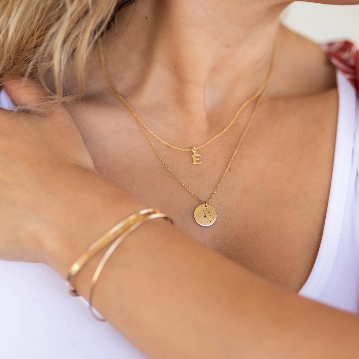 A person wearing two gold chains, one with an initial E pendant and the other with a round constellation charm.