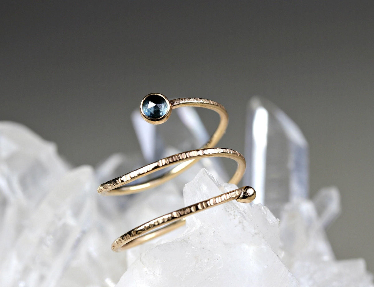 A textured gold wrap ring with a dark blue stone on one end resting on a cluster of raw quartz crystals.